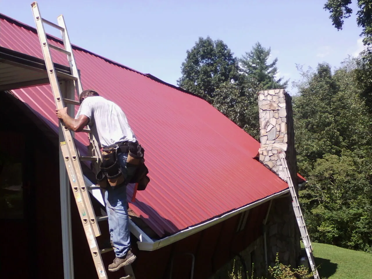 Roofer on a ladder beside a red metal roof and stone chimney — Soffit & Fascia Repair by Roofing Brothers in Cammal